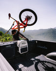 Person loading a bicycle into the back of a pickup truck with mountains in the background