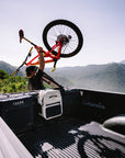 Person loading a bicycle into the back of a pickup truck with mountains in the background