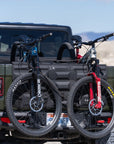 Two bicycles on a bike rack attached to the back of a black SUV with a desert landscape in the background.