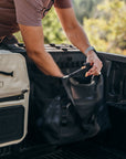 Person loading a black bag into the back of a vehicle with a beige bag beside it.