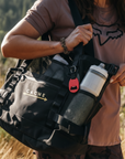 Person holding a black totebag with 'CACHE' branding in a natural setting