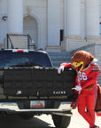 Person in a red and yellow costume leaning on a black truck with a building in the background