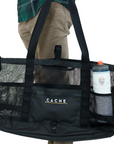 A person holding a black tote bag with the brand 'CACHE' visible on it. The bag features multiple mesh pockets and a water bottle is visible through one of the pockets.