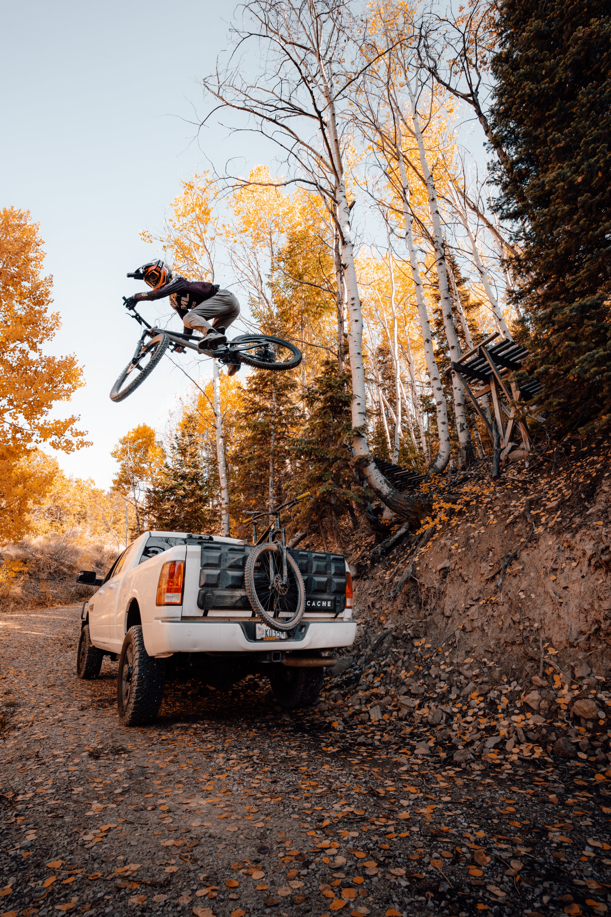 Person on a bicycle in mid-air above a white truck on a dirt road with trees in the background