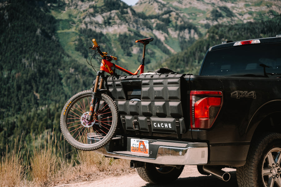 Black truck with a bicycle attached to its bed, set against a mountainous landscape.
