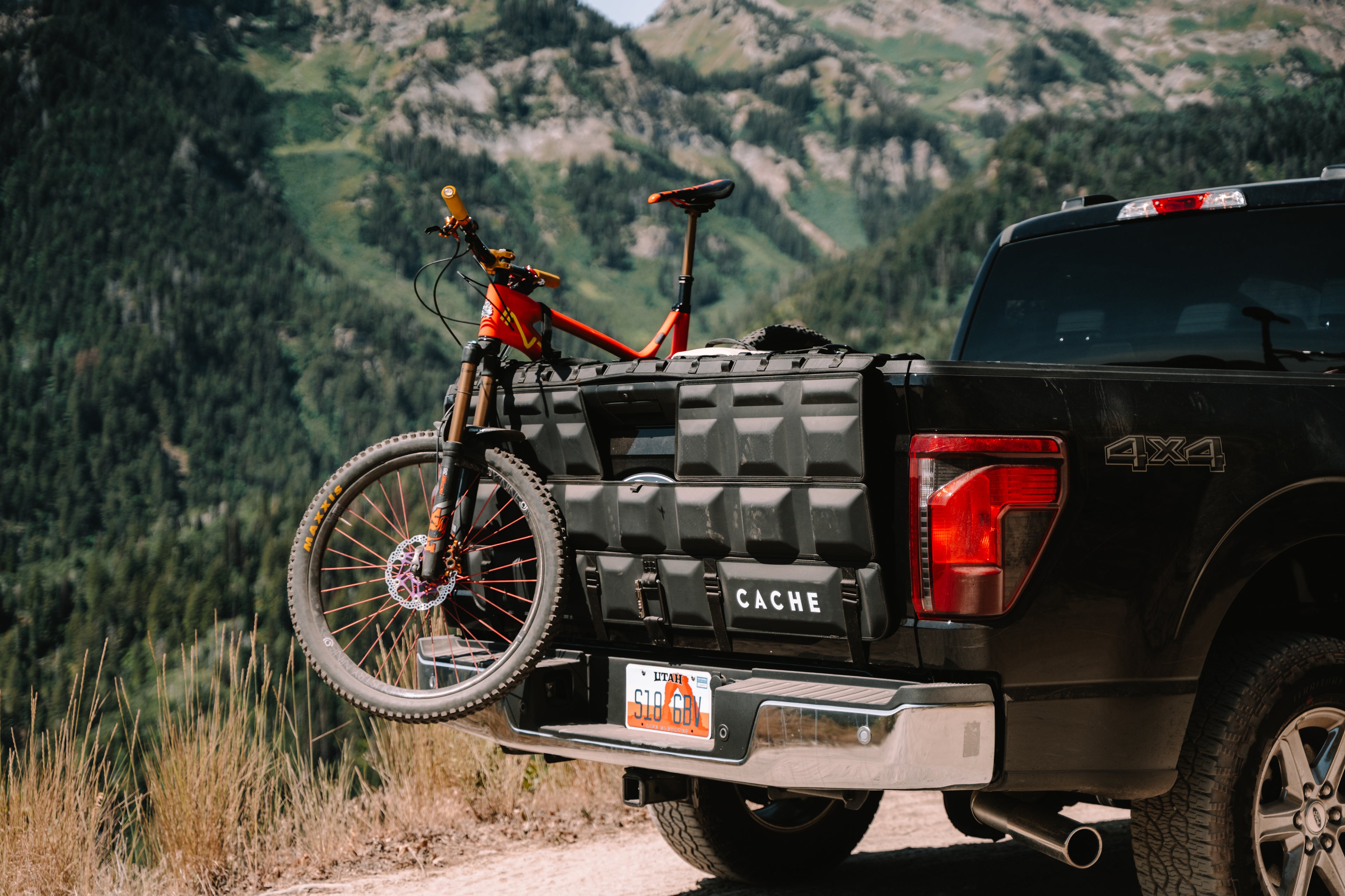 Black truck with a bicycle attached to the back, set against a mountainous landscape.