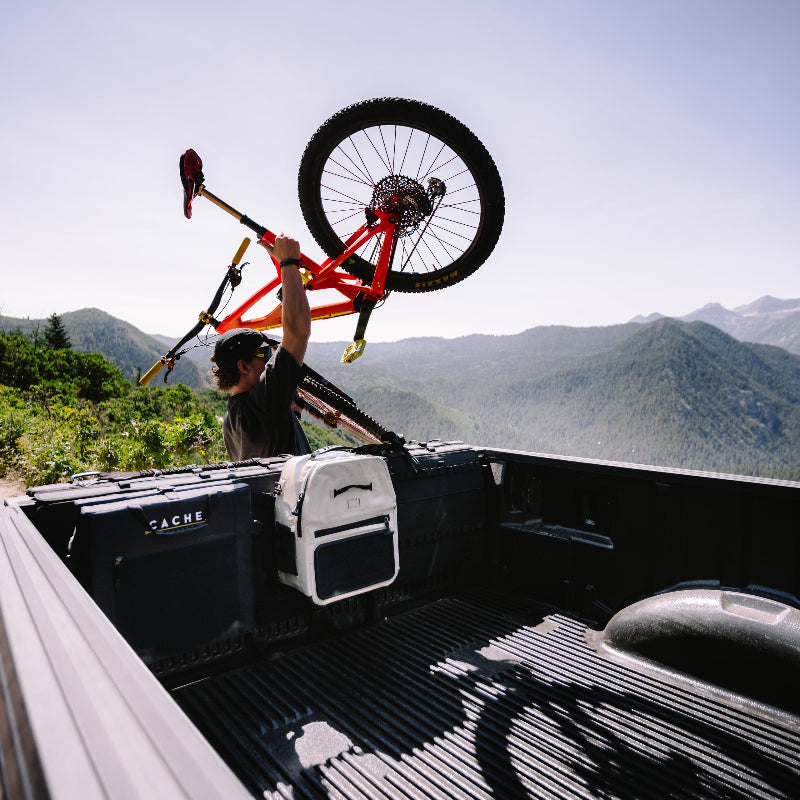 Person loading a bicycle into the back of a pickup truck with mountains in the background