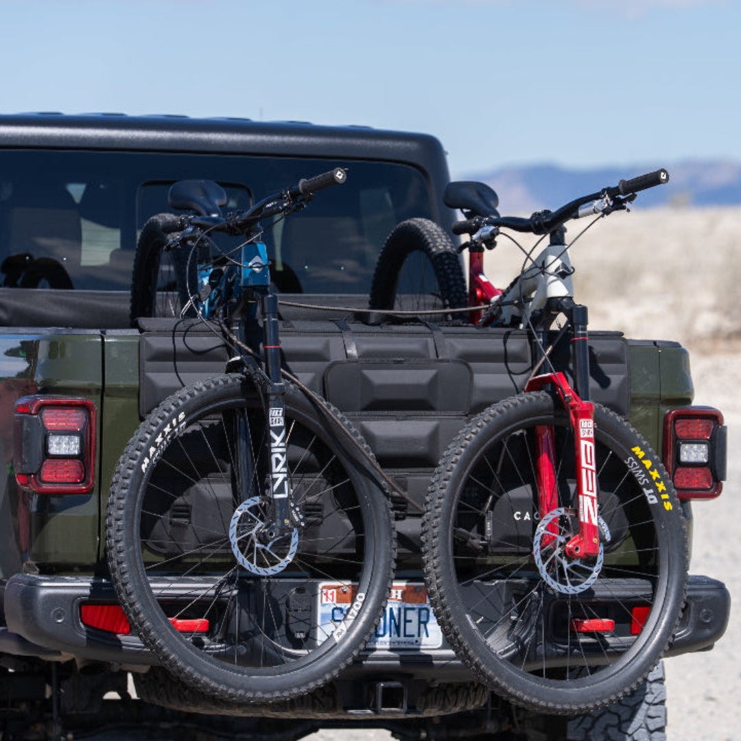 Two bicycles on a bike rack attached to the back of a black SUV with a desert landscape in the background.
