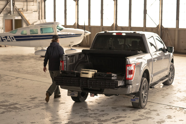 Gray pickup truck with open bed in front of a small airplane inside an airport hangar.