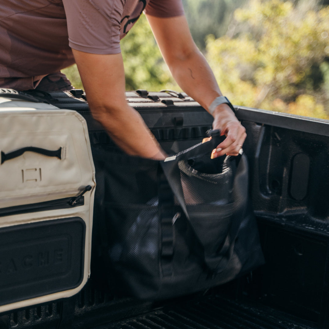 Person loading a black bag into the back of a vehicle with a beige bag beside it.