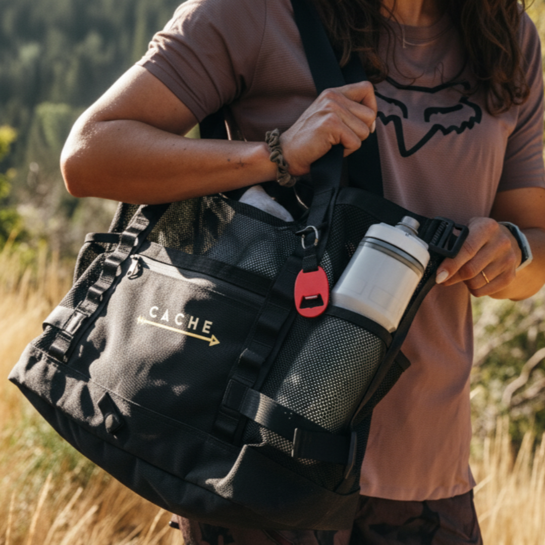 Person holding a black totebag with 'CACHE' branding in a natural setting