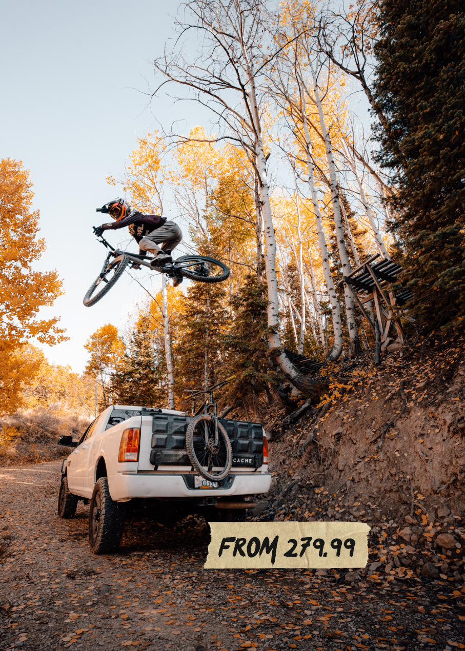 Person on a mountain bike in mid-air above a white truck with bicycles in a forest setting.