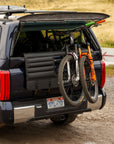 A dark blue truck with a tailgate pad carrying two mountain bikes secured to the back. The truck is parked on a gravel road in a rural setting, with grassy fields and hills in the background. The tailgate pad has straps securing the bikes, and the truck's rear window is open, showing additional bike storage inside