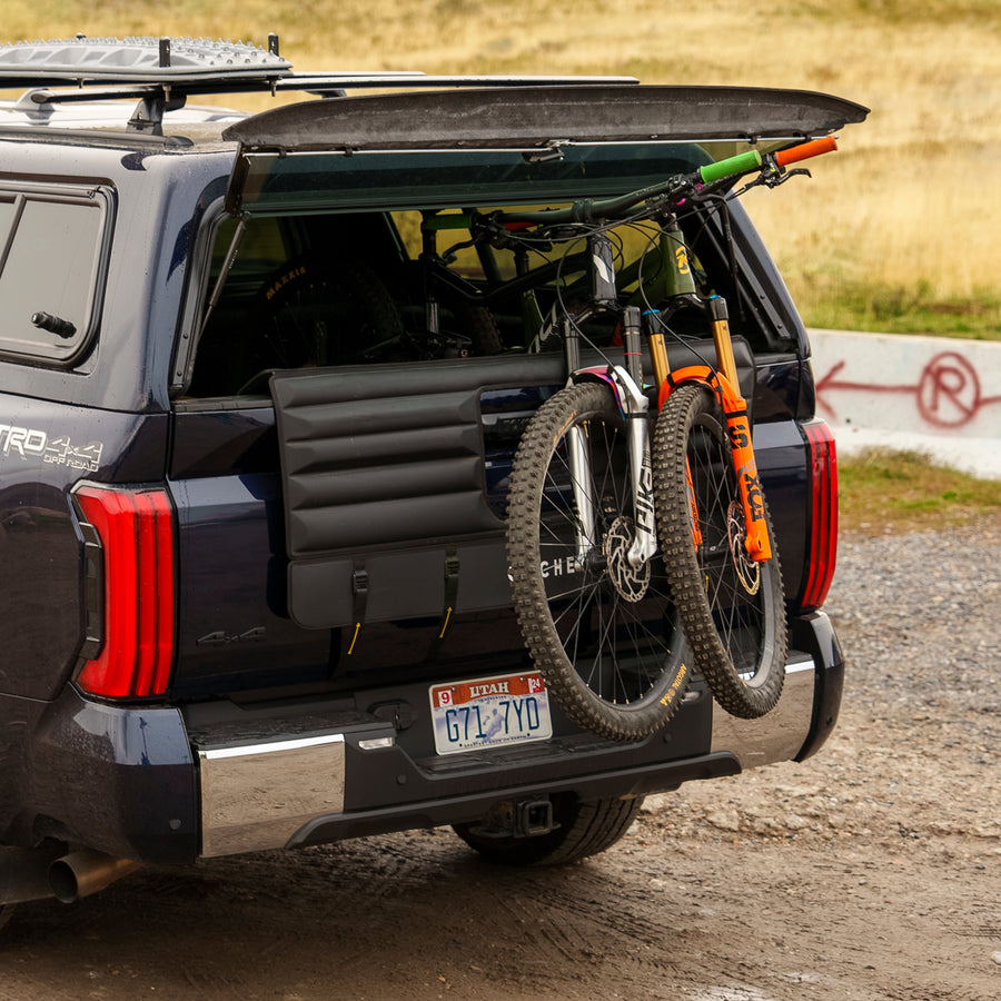 A dark blue truck with a tailgate pad carrying two mountain bikes secured to the back. The truck is parked on a gravel road in a rural setting, with grassy fields and hills in the background. The tailgate pad has straps securing the bikes, and the truck's rear window is open, showing additional bike storage inside