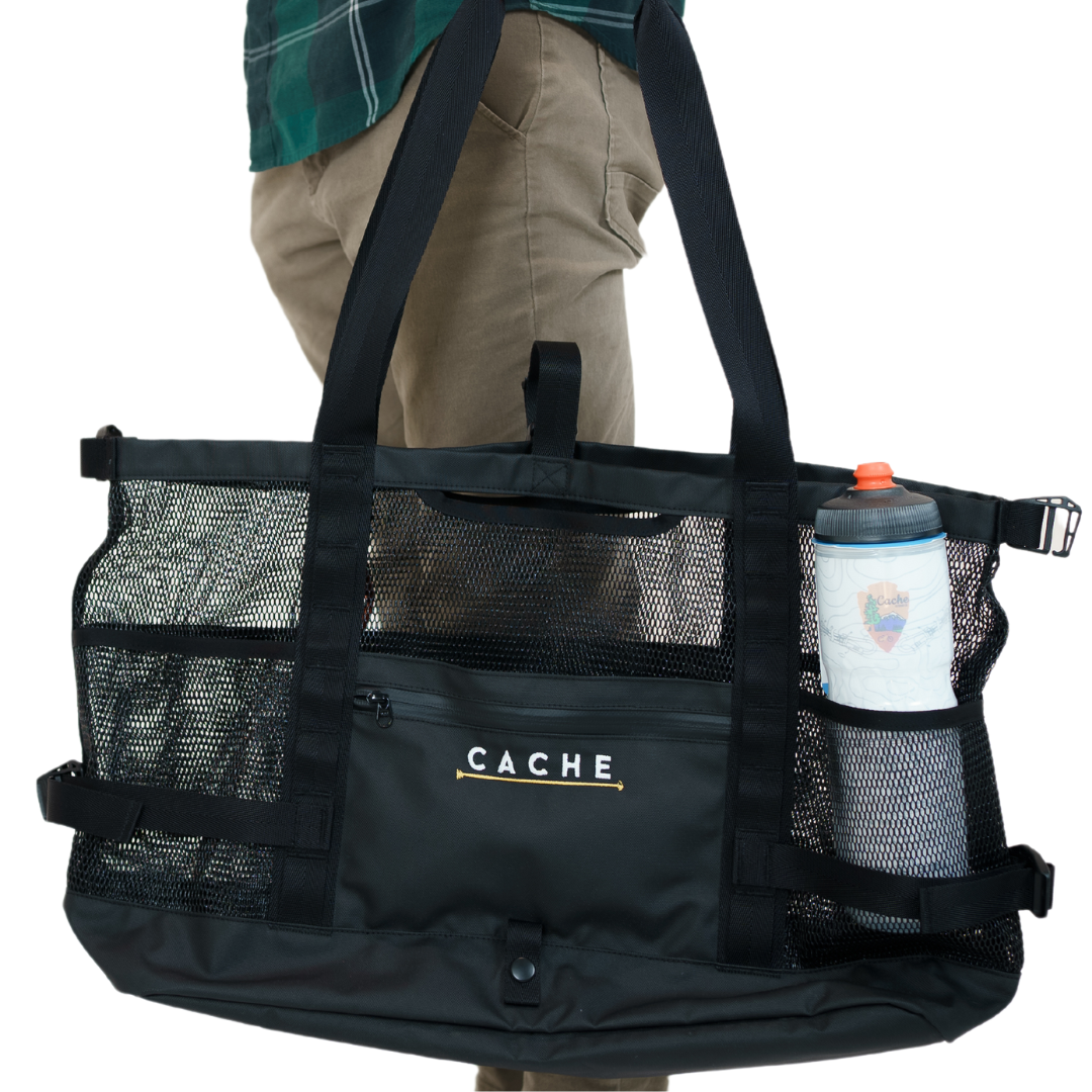 A person holding a black tote bag with the brand 'CACHE' visible on it. The bag features multiple mesh pockets and a water bottle is visible through one of the pockets.